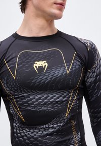 Άνδρας που φοράει μαύρο μακρυμάνικο κομπρεσιονικό μπλουζάκι με μοτίβο φολιδών φιδιού, κίτρινο λογότυπο φιδιού στο στήθος και κίτρινες γραφικές γραμμές.