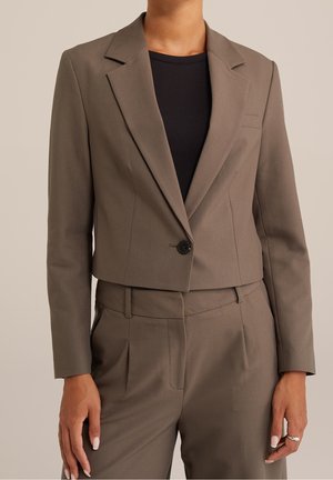 Vrouw met een cropped taupekleurig colbert met één zwarte knoop, gedragen over een zwarte top en bijpassende, hooggetailleerde taupekleurige broek.