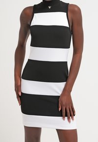 Vestido a rayas en blanco y negro con cuello alto, forma ajustada y textura suave. Destaca un logo en forma de triángulo en el escote.