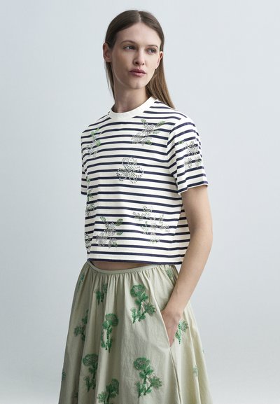 Jeune femme debout les mains dans les poches, portant un haut court rayé avec des imprimés floraux et une jupe beige avec une broderie florale verte.