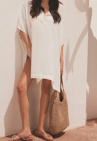 Biała tunika w paski o luźnym kroju, zestawiona z plecioną brązową torbą i beżowymi sandałami z plecionym detalem. Minimalistyczny design.