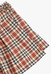 Pleated hame in beige, punainen ja sininen ruudullinen kuosi; vyötäröllä on kaksi kultatukkua; valmistettu tekstuuritetusta kankaasta.