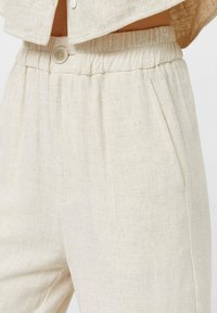 Pantaloni in lino beige con vita elastica, chiusura a bottone e tasche laterali, indossati con un top beige abbinato.