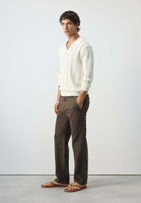 Jeune homme portant un pull en maille blanc à manches longues, un pantalon marron et des sandales beige, debout les mains dans les poches sur un fond clair uni.