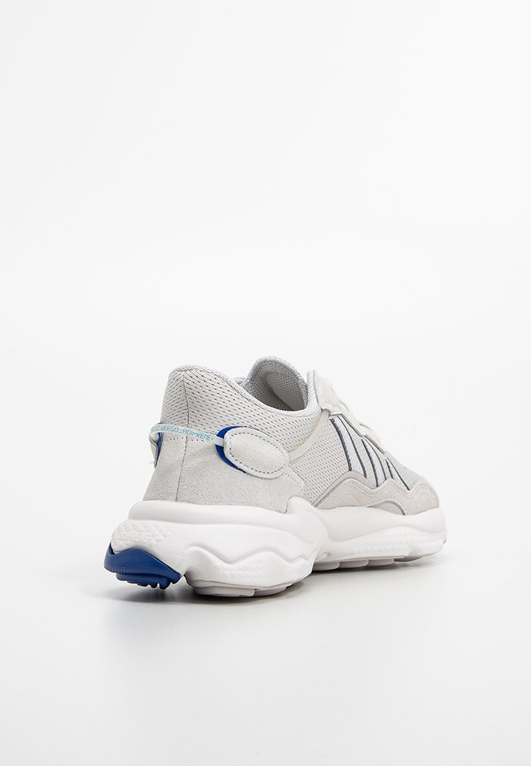 Zapatilla deportiva de color gris claro y blanco con paneles de malla y gamuza, suela gruesa blanca y acento azul en el talón.