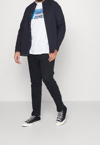 Camicia button-up blu scuro, t-shirt grafica bianca, jeans neri e sneakers nere con suola bianca e dettagli in pizzo. Outfit casual.