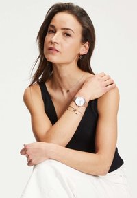 Orologio rotondo bianco con cinturino in metallo rosa dorato, indossato con diversi braccialetti delicati in rosa dorato, caratterizzati da piccoli accenti a forma di cuore.