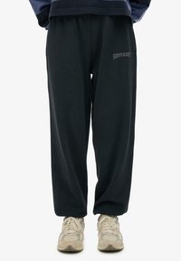 Pantalones de chándal gris oscuro con una cinturilla elástica, bolsillos laterales y logo bordado "SUPERDRY" en gris claro en el muslo izquierdo. Tela texturizada.