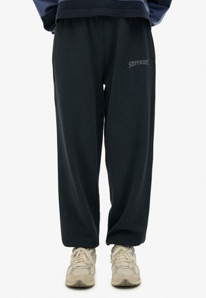Pantalons de survêtement gris foncé avec une taille élastique, des poches latérales et un logo "SUPERDRY" brodé en gris clair sur la cuisse gauche. Tissu texturé.