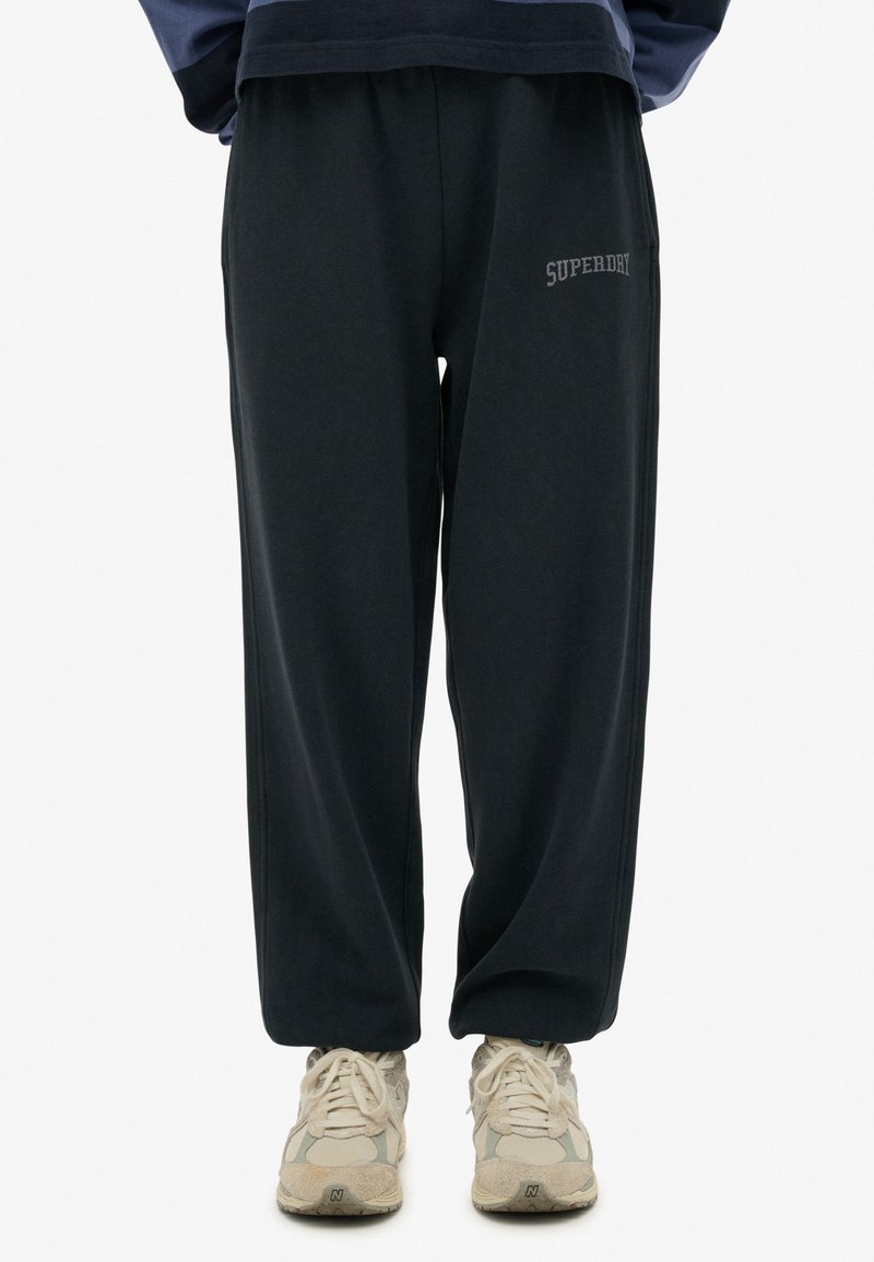 Pantalones de chándal gris oscuro con una cinturilla elástica, bolsillos laterales y logo bordado "SUPERDRY" en gris claro en el muslo izquierdo. Tela texturizada.