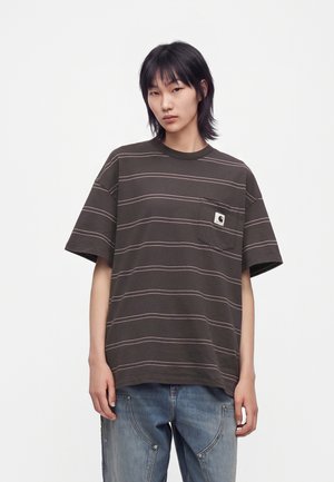 Persona joven con cabello negro lleva una camiseta holgada de rayas marrón oscuro con bolsillo y vaqueros azules sobre un fondo blanco liso.