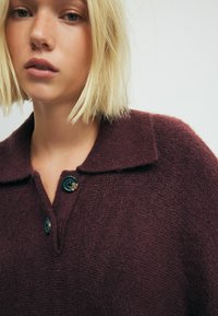 Maronfarvet strikket sweater med klassisk krave, to mørke knapper og en tekstureret overflade. Blødt stof, løs pasform og enkelt design.