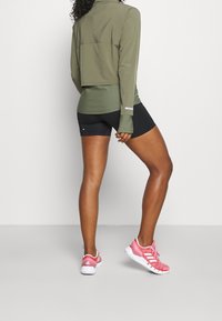 Grön långärmad skjorta med en kortad design, tillsammans med svarta shorts. Sneakers i rosa med vita detaljer. Släta tygtexturer är synliga.