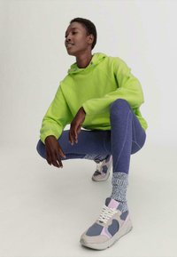 Neon groene hoodie, strakke blauwe legging en multikleurige sneakers met accentdetails. Model poseert in een gehurkte positie.