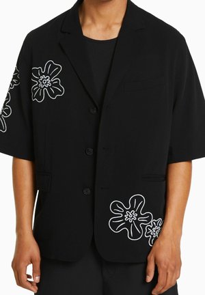 Man draagt een zwart blazer met korte mouwen en witte bloemrijke borduursels op mouw en voorkant, over een zwart overhemd.