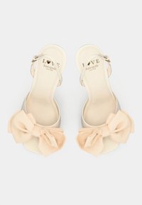 Ανοιχτό μπεζ σανδάλια slingback με μια μεγάλη, μαλακή παστέλ κορδέλα στη μύτη. Λείο υλικό με ελαφριά γυαλάδα και με επενδεδυμένο εσωτερικό.