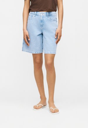 Personne portant un short en jean bleu clair jusqu’aux genoux, un haut marron, des tongs blanches et un bracelet fin à la cheville gauche.
