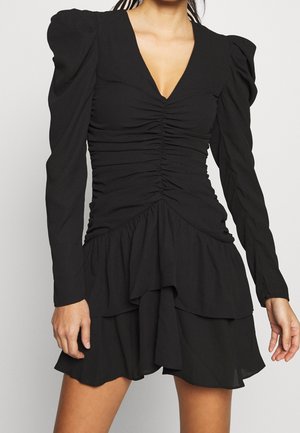 Robe noire à manches longues avec épaules bouffantes, devant froncé, décolleté en V profond et jupe à volants asymétriques superposés portée par une personne debout.