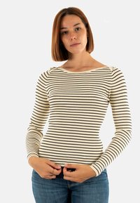 Giovane donna con capelli castani lisci, che indossa una maglietta a righe crema e nera a maniche lunghe e jeans blu, mentre tiene il fondo della sua maglietta.