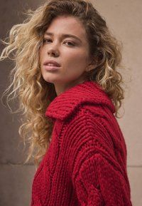 Red knitted sweater with a wide collar, featuring a textured pattern. Model has curly hair, softly illuminated against a neutral background.