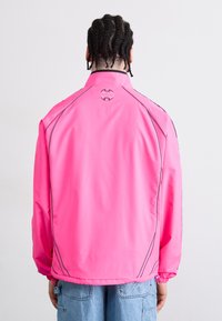 Pink waterproof jacket with a stand collar, elastic cuffs, and a logo detail at the back. Features contrasting stitching and a relaxed fit.