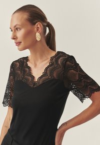 Črni zgornji del z kratkimi rokavi, bogat z izrezom iz čipke, nazobčanim izrezom in gladko tkanino telesa. Videz dopolnjujejo zlate ovalne uhane.