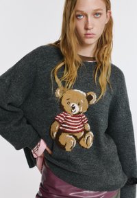 Szary sweter z miękkiego materiału z dzierganym wzorem misia w brązowe i czerwone paski. Luźny krój z ściągaczami na mankietach i u dołu.