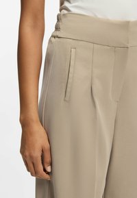 Beige, hoog getailleerde broek met elastische tailleband, uitgevoerd met zijzakken en een gladde, lichte stof. Zichtbare hand rust aan de zijkant.