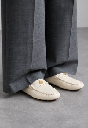 Mocassins en cuir blanc avec emblème doré portés avec un pantalon long gris à jambes larges sur un sol gris.