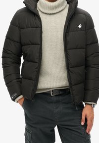 Musta untuvatakki, jossa korkea kaulus, vetoketjullinen etuosa ja erottuva valkoinen logo; yhdistetty vaaleanharmaaseen ribbineulepuseroon ja tummiin housuihin.