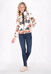 Giacca in pile con design a fantasia arancione, grigio e bianco, con zip frontale e dettagli neri. Abbinata a jeans blu e sneaker beige.