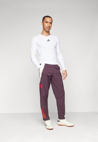 Witte lange mouwen sportshirt met zwart logo, gecombineerd met paarse broek met rode accenten en strepen. Witte sportschoenen met oranje details.