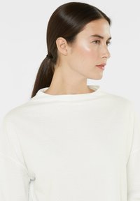 White textured sweater with a high neckline and long sleeves, featuring a subtle woven pattern. Hair is styled in a low ponytail.