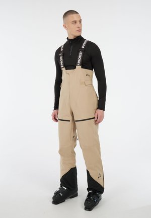 Beige waterdichte skibroek met verstelbare zwarte bretels, zijzakken en contrasterende zwarte onderkanten. Ontworpen voor wintersport.