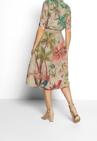 Abito midi a maniche corte in beige con fantasie floreali e di palme colorate. Presenta una vita con cintura ed è abbinato a sandali con tacco a blocco beige.