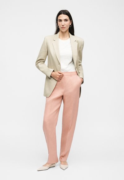 Γυναίκα στέκεται με το ένα χέρι στην τσέπη ροζ ψηλόμεσων παντελονιών, φοράει μπεζ σακάκι, λευκή μπλούζα, κρίκους σκουλαρίκια και μπεζ μυτερές μπαλαρίνες.