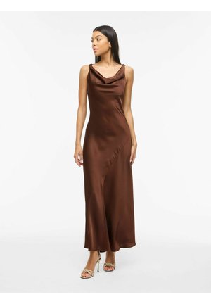 Kvinne iført en ermeløs, brun sateng maxikjole med drapert halslinning og gullflettede høye hæler sandaler, stående mot en hvit bakgrunn.