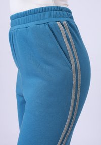 Blå joggebukser laget av mykt stoff, med to sølvstriper på siden og elastisk midje med lommer.