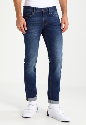 Denim jeans in a dark blue wash with a slim fit, featuring subtle whiskering, five-pocket design, and rolled cuffs. Complemented by white sneakers.