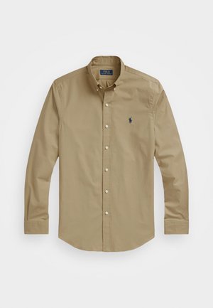 Camicia beige a maniche lunghe con bottoni, colletto abbottonato e piccolo logo scuro sul petto sinistro, distesa su sfondo bianco.