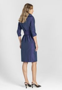 Granatowa sukienka o wykończeniu z kołnierzykiem, przewiązana w talii, z rękawami do trzyczwartych. Gładka faktura, do kolan, o dopasowanej sylwetce.