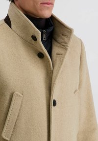 Beige Wollmantel mit hohem Kragen, ausgestattet mit schwarzen Knöpfen und einer darunter sichtbaren, schwarzen Jacke mit Reißverschluss. Strukturiertes Gewebe und sichtbare Nähte.