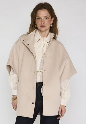 Donna che indossa un cappotto a mantella beige a maniche corte sopra una camicetta bianca di pizzo con fiocco, pantaloni scuri, orecchini a cerchio dorati e diversi anelli e braccialetti d'oro.