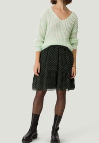 Pull en tricot vert clair avec un col en V, associé à une jupe noire ornée d'un motif floral doré et d'un ourlet volanté, et des bottines noires.
