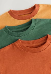 Trois t-shirts à col rond : un orange, un vert et un jaune moutarde. Tous présentent des cols côtelés et un tissu texturé.