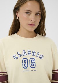 Sudadera color crema con estampado azul "CLASSIC 86", que cuenta con mangas de rayas burdeos y un cuello acanalado. Material de algodón suave.