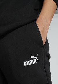 Mustad mustadest dressipükstest pehmest kangast, millel on vasaku tasku peal valge Puma logo, tavalise lõikega ja küljetaskute detailidega.