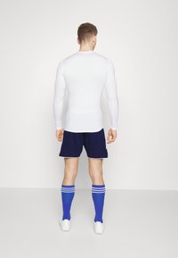 Witte lange mouwen top, marineblauwe shorts, blauwe kniekousen met witte strepen en witte voetbalschoenen. Soepele stof, strakke pasvorm, atletisch ontwerp.
