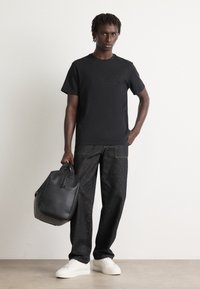 Man in zwart t-shirt en jeans, die een zwarte leren sporttas vasthoudt, staand op een betonvloer tegen een witte muur.