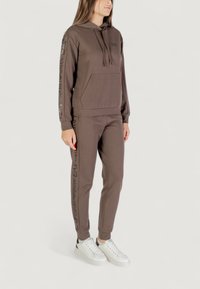 Mujer vistiendo una sudadera con capucha marrón y joggers a juego con los logotipos de EA7 Emporio Armani en las mangas y en las piernas del pantalón, combinados con zapatillas blancas.